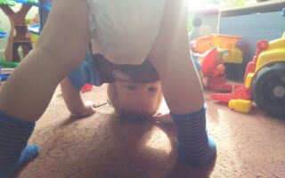 A toddler wearing a nappy and striped socks bends over, looking upside down between their legs in a playroom filled with colourful toys and sunlight streaming in from behind.