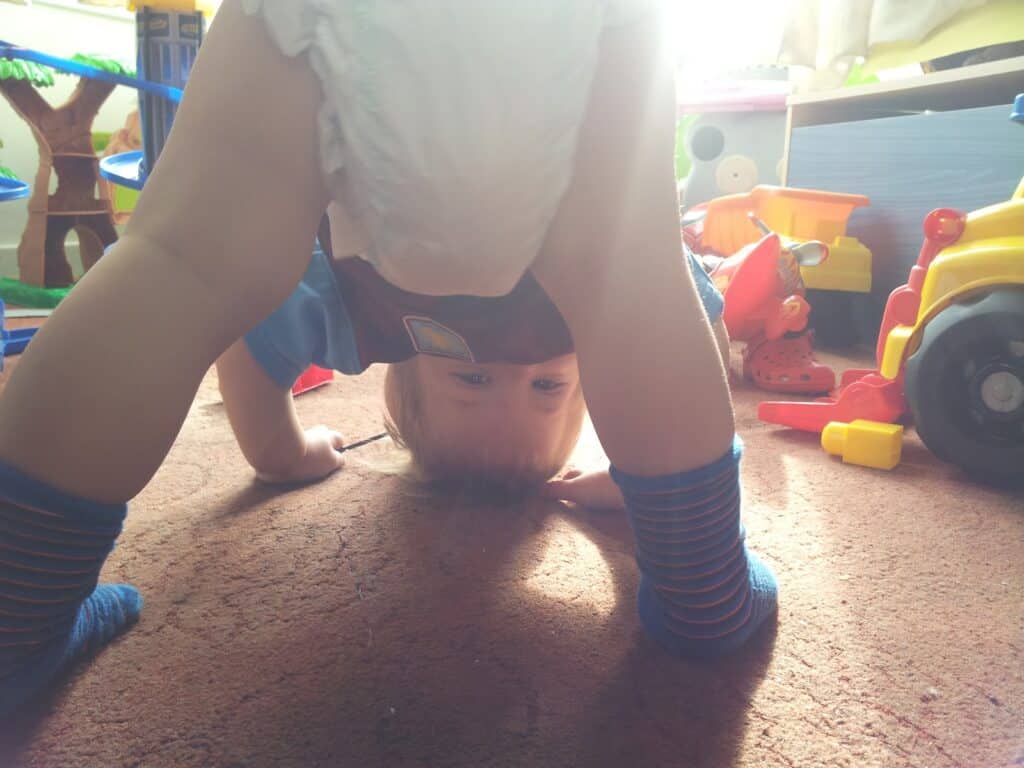 A baby in a nappy and striped socks bends over, looking upside down between their legs in a sunlit playroom, surrounded by colourful toys and a large yellow toy vehicle on a brown carpet. Sunlight streams in from behind the baby.
