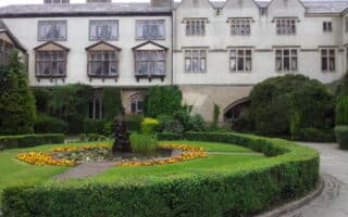 An historic stone building with many windows overlooks a formal garden. The garden features a circular lawn with a central flower bed of yellow and orange flowers, a small fountain or statue, and is surrounded by manicured hedges and greenery.