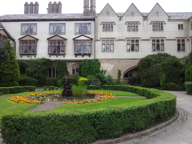 An historic stone building with many windows overlooks a formal garden. The garden features a circular lawn with a central flower bed of yellow and orange flowers, a small fountain or statue, and is surrounded by manicured hedges and greenery.