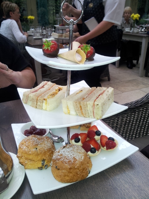 A three-tiered afternoon tea stand holds cakes, sandwiches, and scones. The top tier has chocolate-covered strawberries and a rolled pastry, the middle tier has assorted finger sandwiches, and the bottom tier features scones with berries and fruit-topped tartlets.