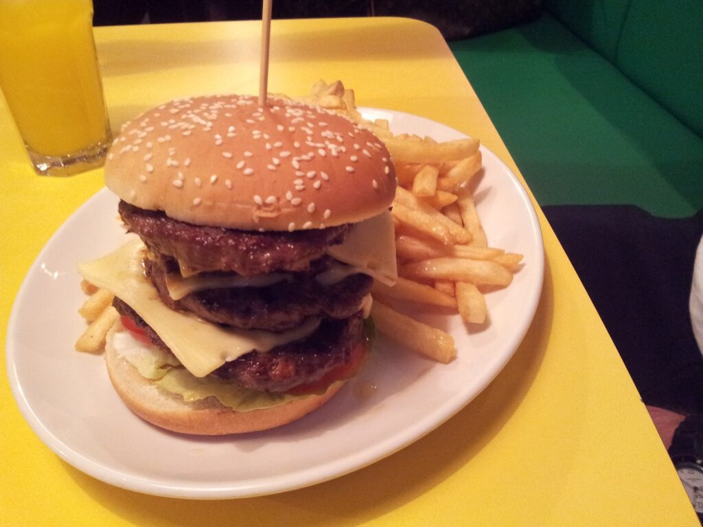A large hamburger with three beef patties, several slices of cheese, lettuce, and tomato is stacked high on a sesame seed bun. It is served on a white plate with a generous portion of chips. A glass of orange drink is visible in the background.