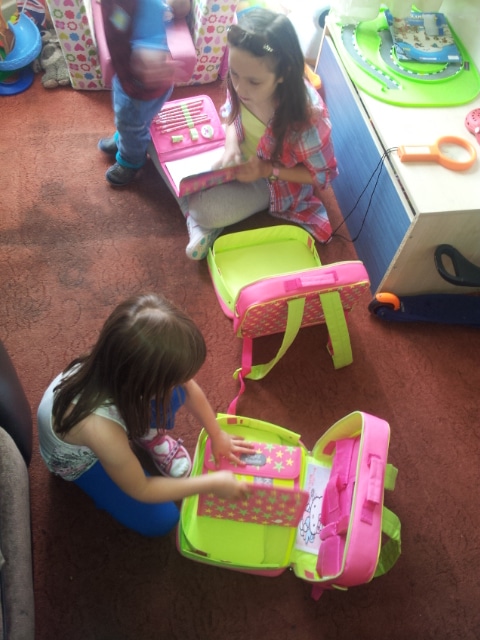 Two young children sit on a carpeted floor playing with pink and green toy make-up kits. One child wears a white top and blue trousers, while the other has a checked shirt and headband. A third child stands nearby. A toy doctor’s kit is on a table beside them.