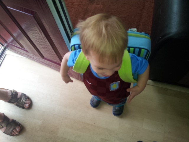 A young child with light brown hair wearing a maroon and blue sports kit and blue trainers stands indoors. The child has a green-strapped blue rucksack and looks slightly downward. The child is near an open door; an adult’s sandals are visible on the left.