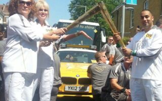 Three torchbearers in white uniforms hold Olympic torches together in front of a yellow bus with “Moment to shine” displayed. Supporters and staff in grey gather around, with a clear blue sky and brick buildings in the background.