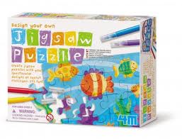 A box for a “Design Your Own Jigsaw Puzzle” kit. The front shows completed puzzle pieces with colourful fish and sea creatures. The packaging includes bright lettering and art supplies, with branding and age recommendations in the lower corners.