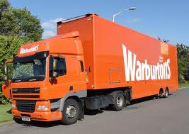 A large orange Warburtons lorry is parked on a road by greenery. The cab and trailer both feature the white Warburtons logo. The sky is clear and a streetlamp is visible above the trailer.