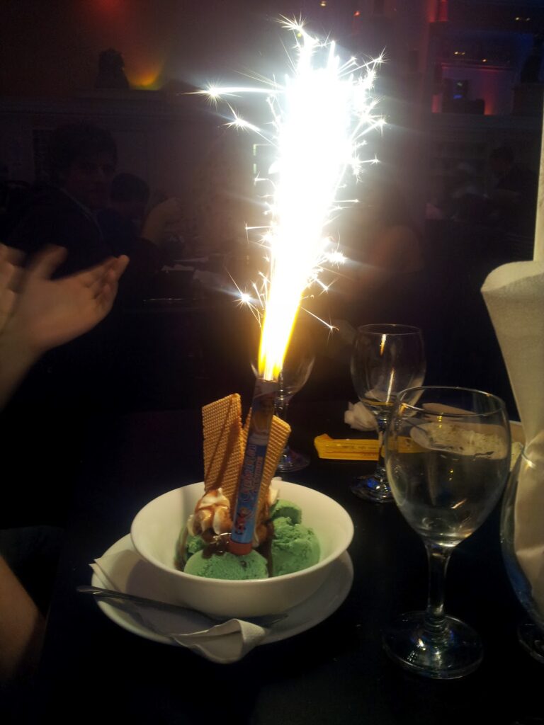 A bowl of green ice cream with wafer biscuits and whipped cream has a lit sparkler on top, emitting bright sparks. There are empty wine glasses on the table, and a person's hands are visible, clapping nearby in a dimly lit restaurant.