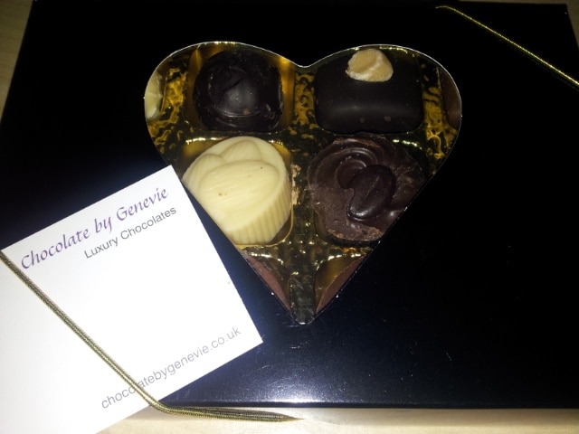A heart-shaped box containing four assorted luxury chocolates sits on a black surface. A white card labelled "Chocolate by Genevie Luxury Chocolates" and a gold ribbon lie beside the box. The chocolates are various shapes and colours.