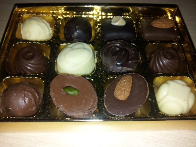 A gold box containing twelve assorted chocolates, including milk, dark, and white varieties. Some chocolates are round or square, and a few are topped with nuts like almonds or pistachios. The chocolates are arranged in neat rows in a plastic tray.