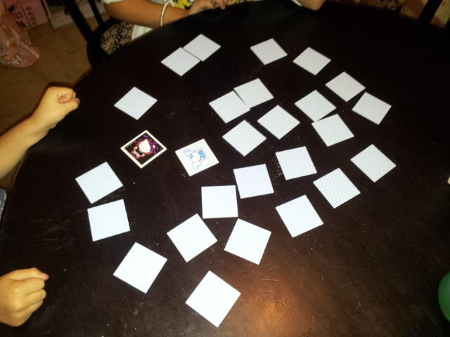 Three children’s hands are visible around a black table with a memory card game in progress. Several cards are face down whilst two cards are face up, showing cartoon images. The children’s fists suggest anticipation or excitement.