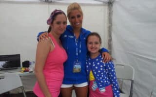 Three people stand together inside a white marquee. An adult woman in a pink vest and headband is on the left. In the centre, a woman in a blue jacket with a media badge has her arms around the others. On the right is a smiling girl in a blue spotty jumper.