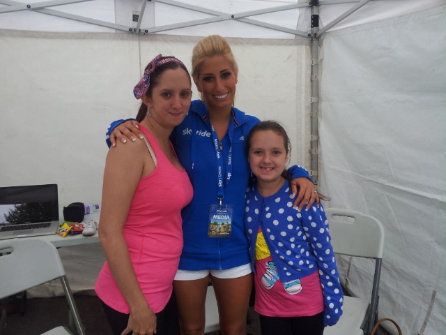 Three people stand together inside a white marquee. An adult woman in a pink vest and headband is on the left. In the centre, a woman in a blue jacket with a media badge has her arms around the others. On the right is a smiling girl in a blue spotty jumper.