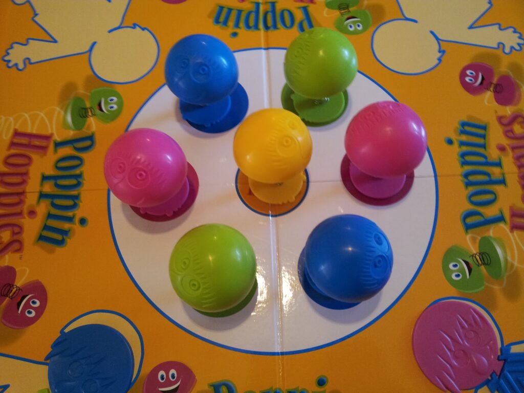 A close-up of a colourful board game called "Poppin Hoppies" shows seven plastic, round figures in blue, pink, green, and yellow standing upright in a white circle at the centre of the bright, cartoon-filled game board.