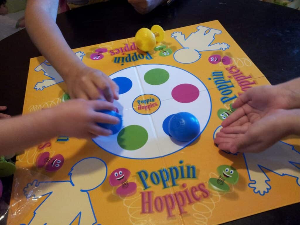 Three children's hands play the game Poppin Hoppies. The colourful board features cartoon figures and circles, with blue, yellow, and green spring-loaded toys on it. One hand is open, ready to catch a toy, while others reach for pieces.