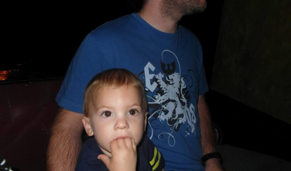 A young child with light brown hair sits in front of an adult man wearing a blue graphic T-shirt. The child looks ahead with fingers in their mouth, while the man, whose face is partially out of frame, looks to the side. The background is dark and indistinct.