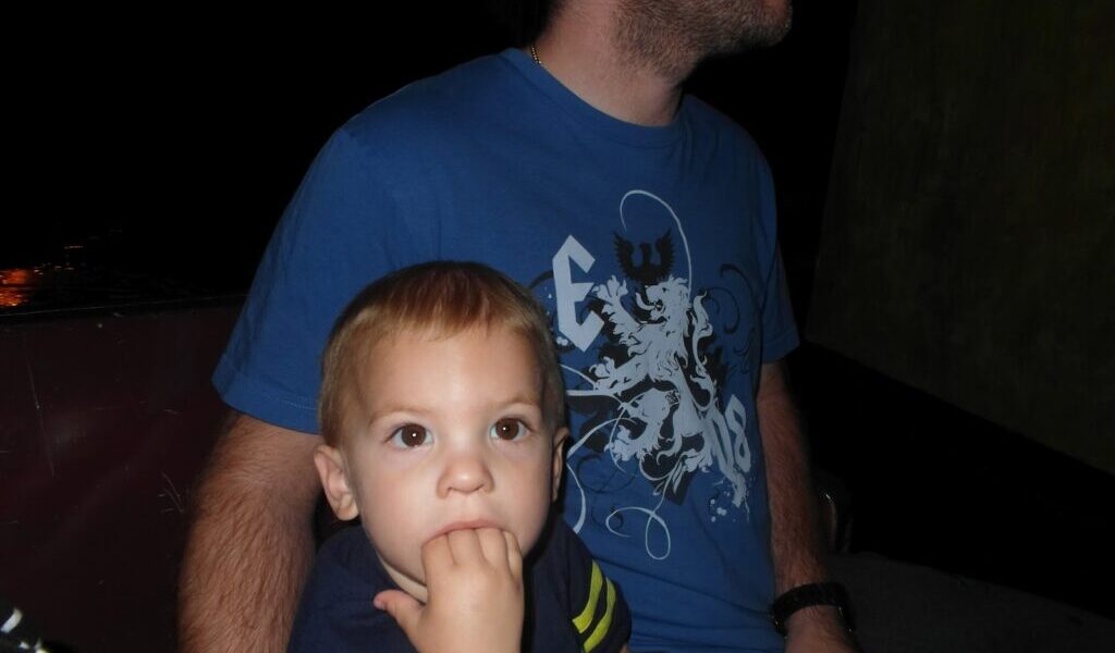 A young child with light brown hair sits in front of an adult man wearing a blue graphic T-shirt. The child looks ahead with fingers in their mouth, while the man, whose face is partially out of frame, looks to the side. The background is dark and indistinct.