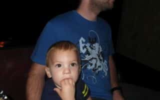 A young child with light brown hair sits in front of an adult man wearing a blue graphic T-shirt. The child looks ahead with fingers in their mouth, while the man, whose face is partially out of frame, looks to the side. The background is dark and indistinct.