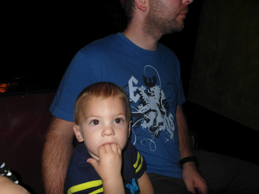 A young child with short light brown hair sits on an adult's lap, looking slightly to the side with fingers in their mouth. The adult, wearing a blue graphic T-shirt, is partially out of frame and faces sideways. The background is dark and indistinct.