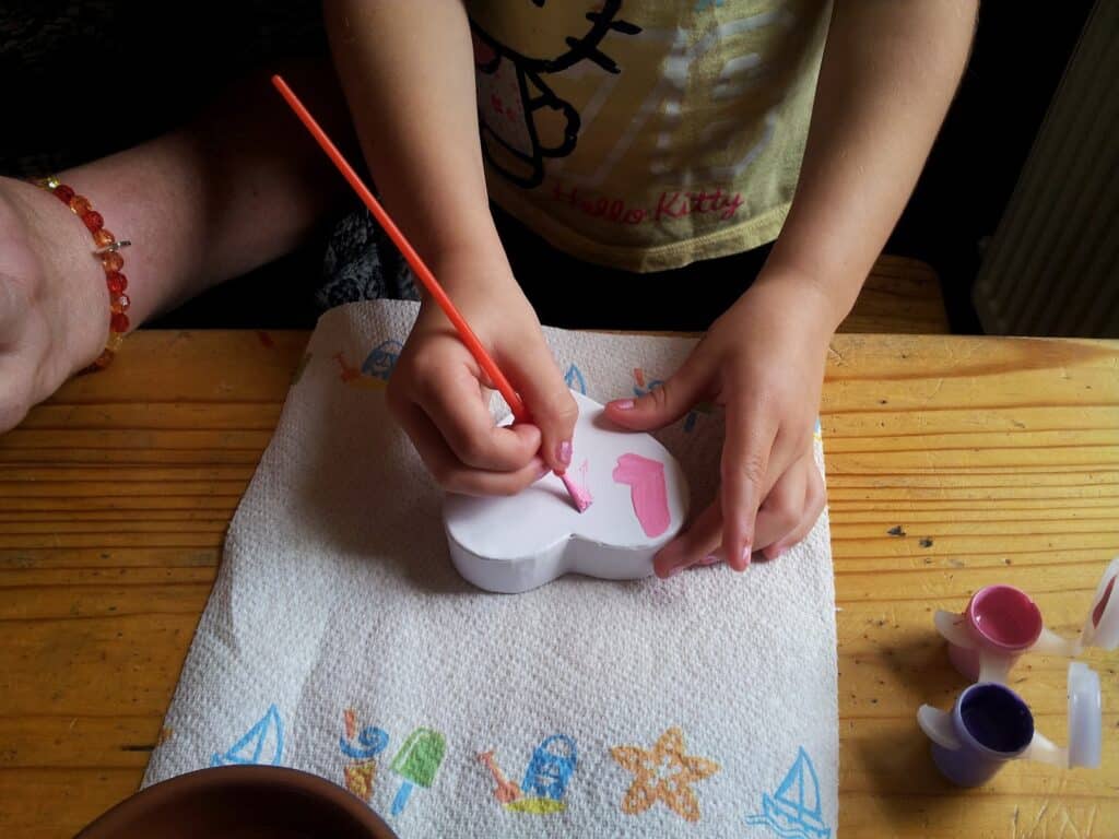 A child in a yellow Hello Kitty shirt paints a white, heart-shaped object pink using a brush. The object rests on a paper towel decorated with colourful beach-themed drawings. Several small paint pots and a hand are visible on the wooden table.