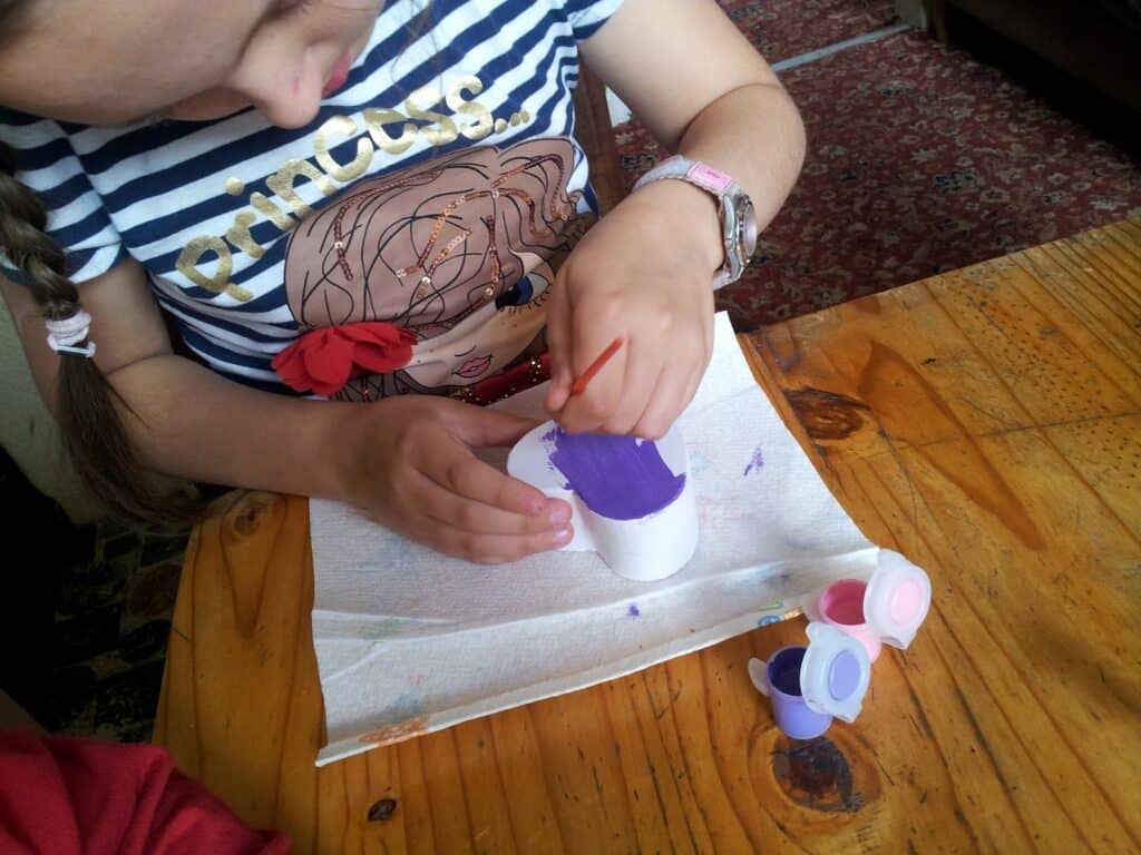 A child wearing a striped "Princess" shirt and a pink watch paints a small white object purple with a brush. The object sits on a kitchen roll, and containers of pink and purple paint are nearby on a wooden table. The child's face is partially out of shot.