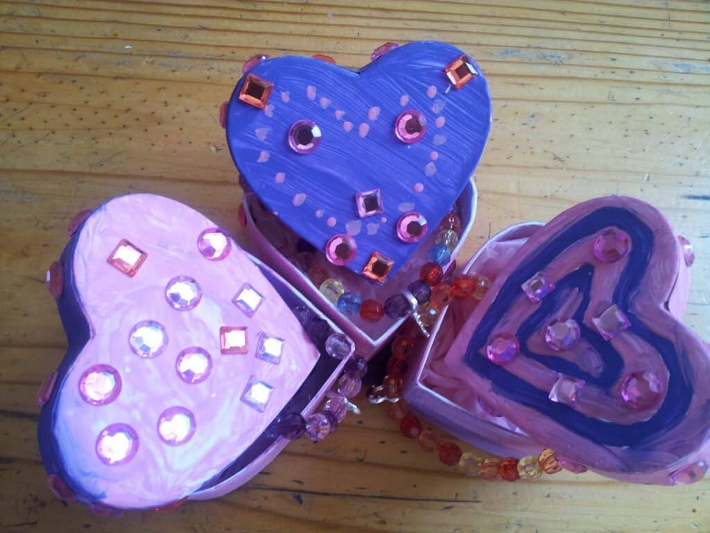Three small, heart-shaped boxes painted pink, purple, and light purple sit on a wooden surface. Each lid is decorated with shiny sequins and beads, creating sparkling patterns. Beaded strings are visible, adding colourful accents to the arrangement.
