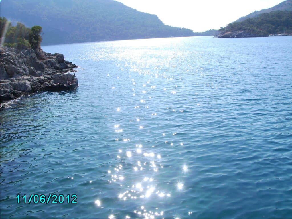 A bright, sunny day over a calm blue sea with sparkling reflections. Rocky shores and green hills frame the water. The date "11/06/2012" is displayed in turquoise text in the lower left corner. The sky is clear and hazy in the distance.