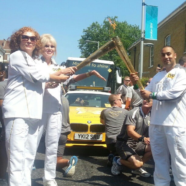 Three torchbearers in white uniforms are passing the Olympic flame between gold torches on a sunny street, with a yellow Olympic support vehicle and cheering people in grey shirts behind them. A blue Olympic banner hangs on a streetlamp.
