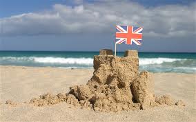 A small sandcastle sits on a sandy beach, adorned with a miniature Union Jack flag on top. In the background, gentle sea waves roll under a partly cloudy sky. The scene suggests a playful, summery day by the seaside.