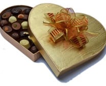 A heart-shaped golden box, partially opened to reveal an assortment of chocolates inside. The lid, decorated with an orange and gold ribbon bow, rests beside the chocolates. The background is white.
