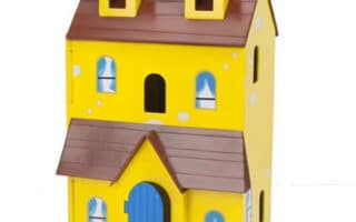 A yellow, two-storey toy house with a brown roof and two dormer windows. It has a blue arched front door, four windows with blue curtains on the ground floor, and three windows on the first floor. The house has a whimsical, cartoon-like design.