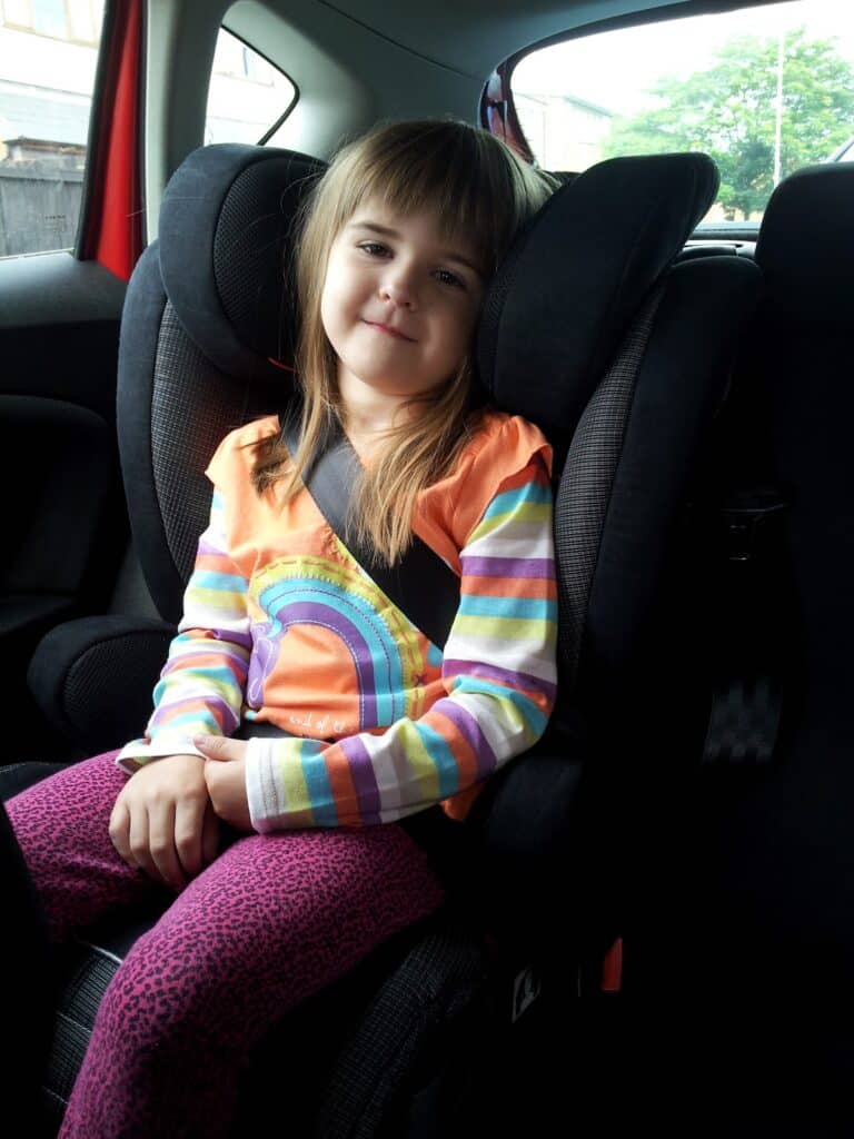 A young child with straight, light brown hair sits in a car seat, secured by a seatbelt. She wears a colourful striped shirt with a rainbow design and purple patterned trousers, smiling gently whilst looking towards the camera. The car interior is visible around her.