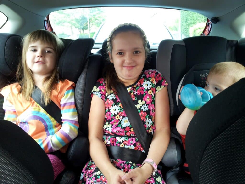 Three children sit in car seats in the back of a car. The girl on the left wears a rainbow-striped top, the girl in the middle wears a floral dress and smiles with hands clasped, and the toddler on the right drinks from a blue beaker. All wear seatbelts.