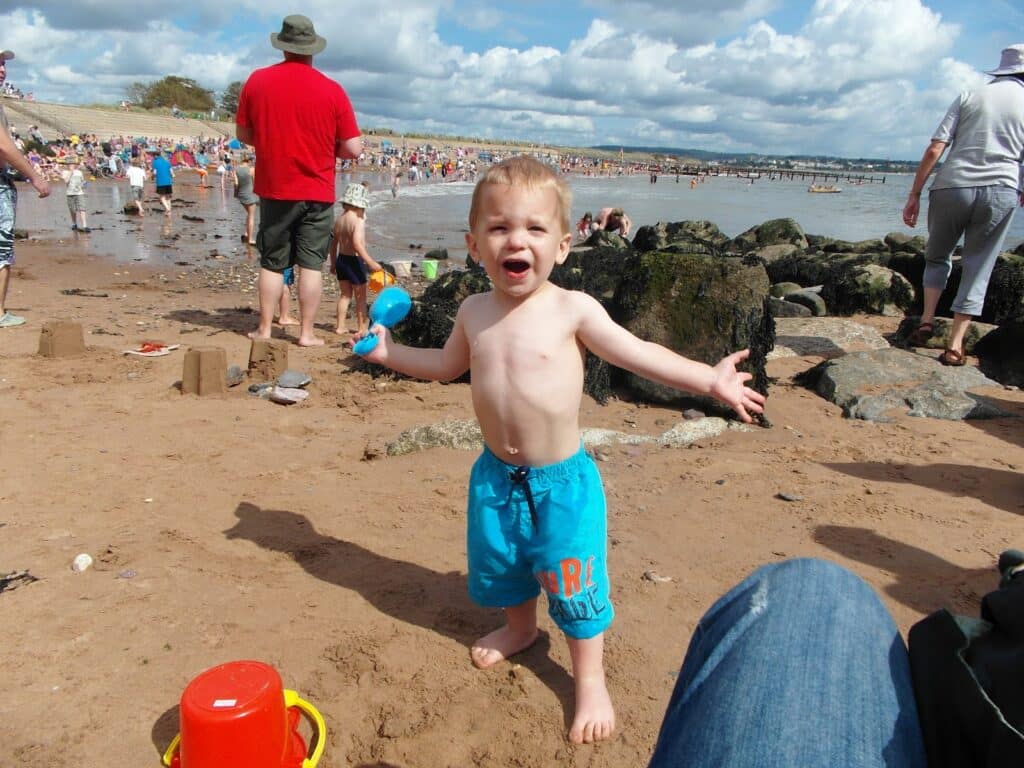 A young boy in blue swimming shorts stands barefoot on a sandy beach, arms outstretched and smiling. He holds a blue toy spade. People, rocks, and sandcastles are in the background, with the sea, a cloudy sky, and crowds enjoying the sunny day at the seaside.