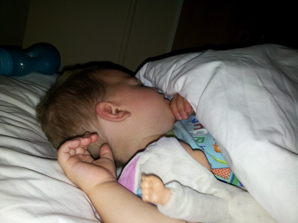 A baby is sleeping on a bed, partially covered by a white blanket. The baby is lying on its side, with one hand near its face and the other arm holding a soft toy. A blue beaker is visible in the background on the bed.