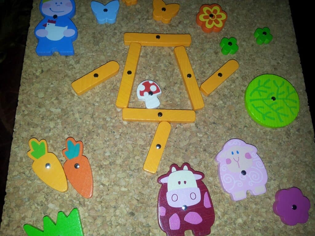A corkboard displays colourful wooden pieces: animals, carrots, butterflies, flowers, a green circle, and orange sticks forming a frame with a mushroom inside. A blue figure, pink sheep, purple cow, and leaves are also included among the playful shapes.