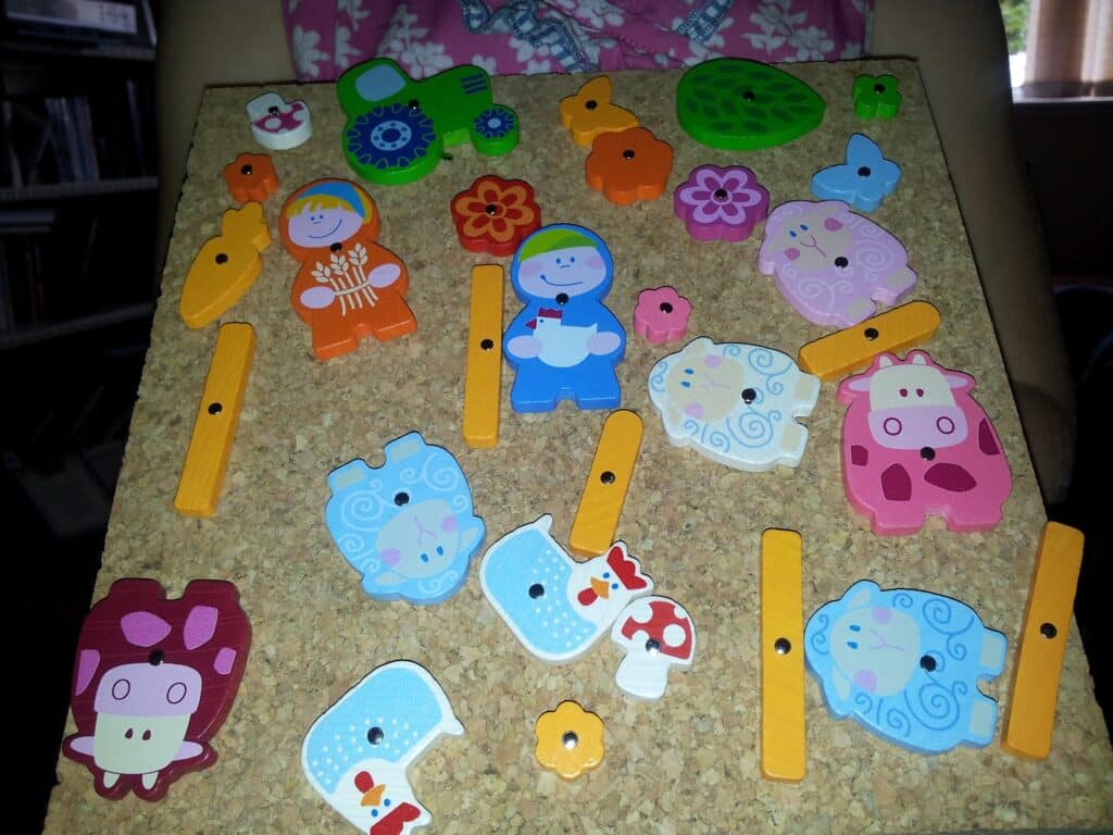 A cork board displays colourful animal and character pieces, flowers, and mushrooms, fastened with yellow pegs. Visible pieces include cows, sheep, pigs, and children in fancy dress. The background shows a person holding the board against their body.