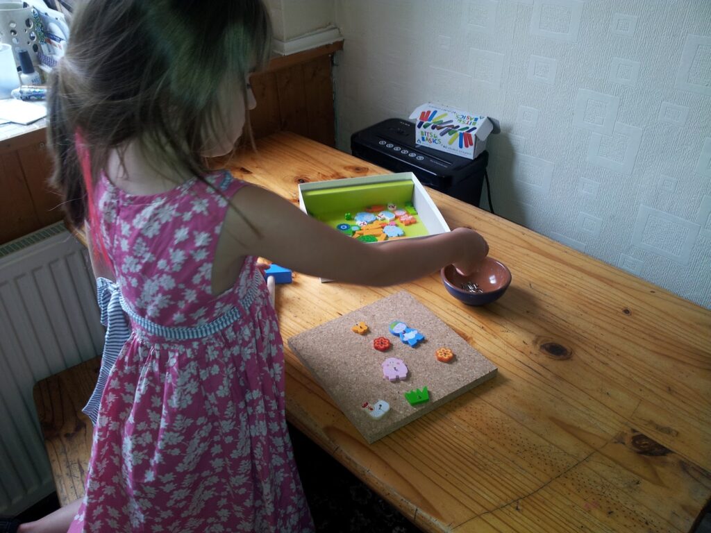 A young girl in a pink floral dress arranges colourful buttons and shapes on a corkboard, which sits on a wooden table. A small brown bowl and a yellow tray holding more buttons are nearby, and a printer is in the background.