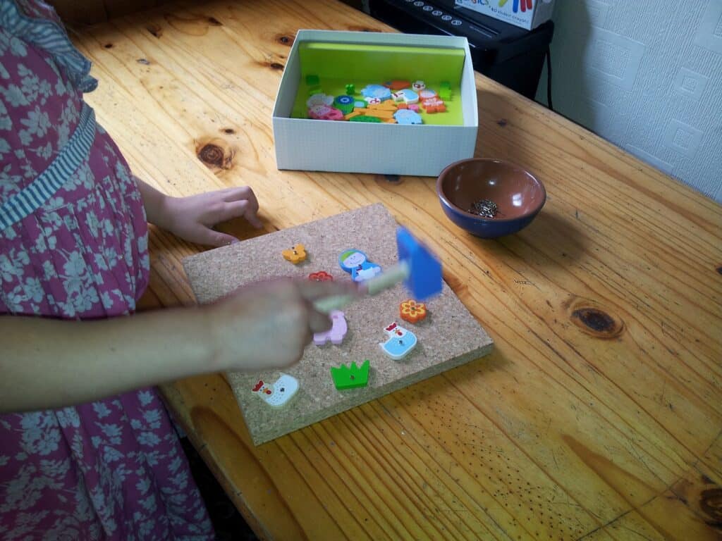 A child in a pink patterned dress uses a small blue hammer on a corkboard with colourful animal and object shapes. A box with shapes and a brown bowl are on the wooden table. Only the child’s arms and torso are visible.