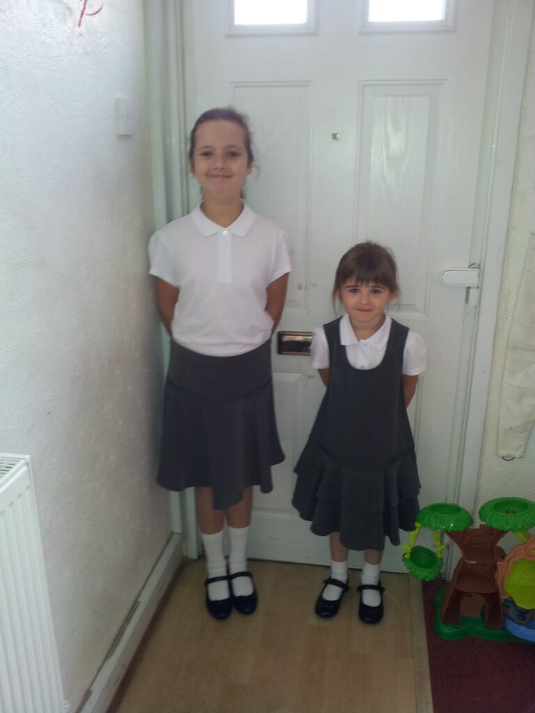 Two young girls stand side by side in front of a white door. Both wear school uniforms: white shirts, grey skirts or pinafores, white socks, and black shoes. The older girl smiles, hands behind her back; the younger girl stands with hands at her sides.