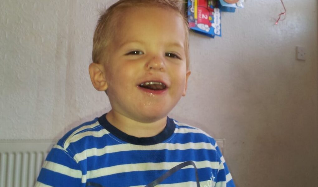 A young child with short blond hair and a happy expression is wearing a blue and white striped shirt. The child is sitting indoors with a cream-coloured textured wall and some colourful hanging decorations in the background.