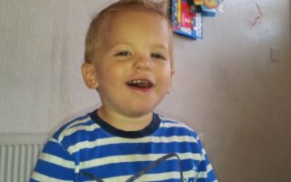 A young child with short blond hair and a happy expression is wearing a blue and white striped shirt. The child is sitting indoors with a cream-coloured textured wall and some colourful hanging decorations in the background.