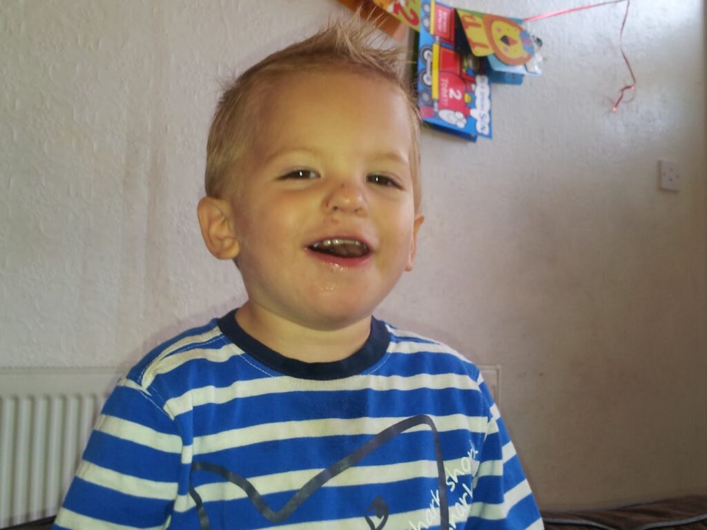 A young child with short blond hair smiles, wearing a blue and white striped shirt. The background shows a cream-coloured wall, a white radiator, and colourful decorations or packages hanging near the top right. The lighting is natural.
