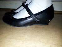 A close-up side view of a child's feet wearing white socks and black patent leather Mary Jane shoes with a strap, standing on a light wooden floor.