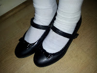 A close-up of a child's feet wearing white socks and shiny black Mary Jane shoes with a small bow on each toe, standing on a light-coloured wooden floor.