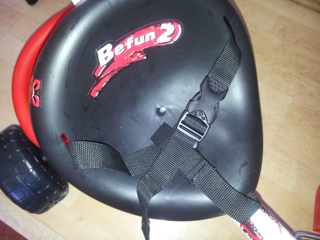 Close-up of a black plastic bicycle or tricycle seat labelled "Befun 2" in red and white letters, with black safety straps attached. The seat is set against a light wooden floor, and part of a wheel and red frame are visible in the background.