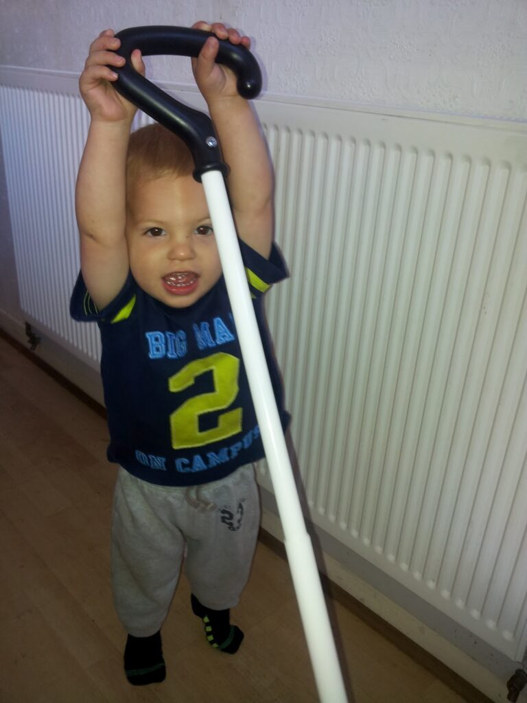 A smiling toddler stands indoors, holding a white cane with a black curved handle above their head. The child wears a navy shirt with a large yellow "2" and grey tracksuit bottoms. The background features a white radiator and wooden floor.