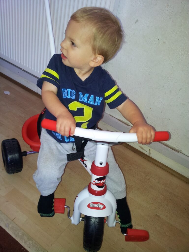 A toddler with fair hair, wearing a navy shirt reading “BIG MAN 2” and grey tracksuit bottoms, sits on a white and red Smoby tricycle indoors. The child looks to the left, holding onto the handlebars with both hands. The background shows a radiator and wall.