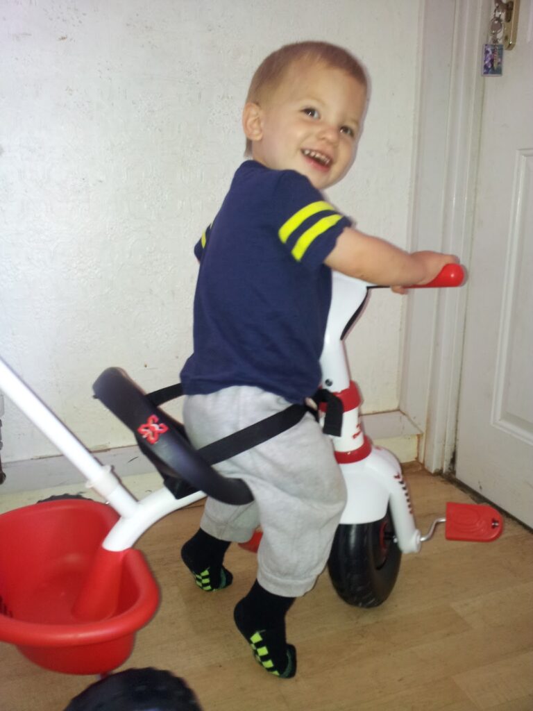 A smiling toddler in a navy blue shirt with yellow stripes and light grey trousers sits on a white and red tricycle indoors. The child is wearing black socks with green accents and is looking back over one shoulder, appearing happy and playful.
