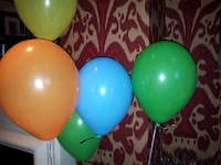 Five balloons—orange, yellow, blue, and two green—are grouped together indoors in front of red-and-beige patterned wallpaper. The balloons appear shiny and are tied with strings, some floating higher than others.